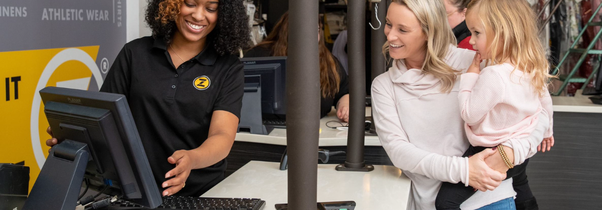 A female employee behind a checkout counter at ZIPS cleaners speaks with a woman across the counter who is holding a child.