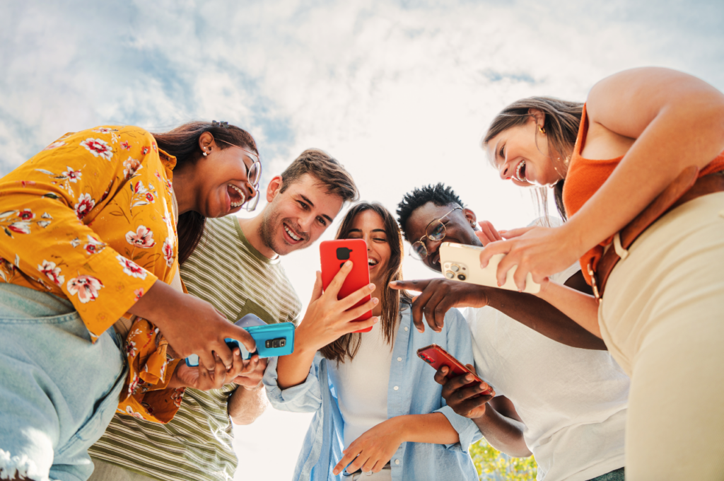 A group of five adults stand looking at their mobile phones in their hands, smiling and laughing.