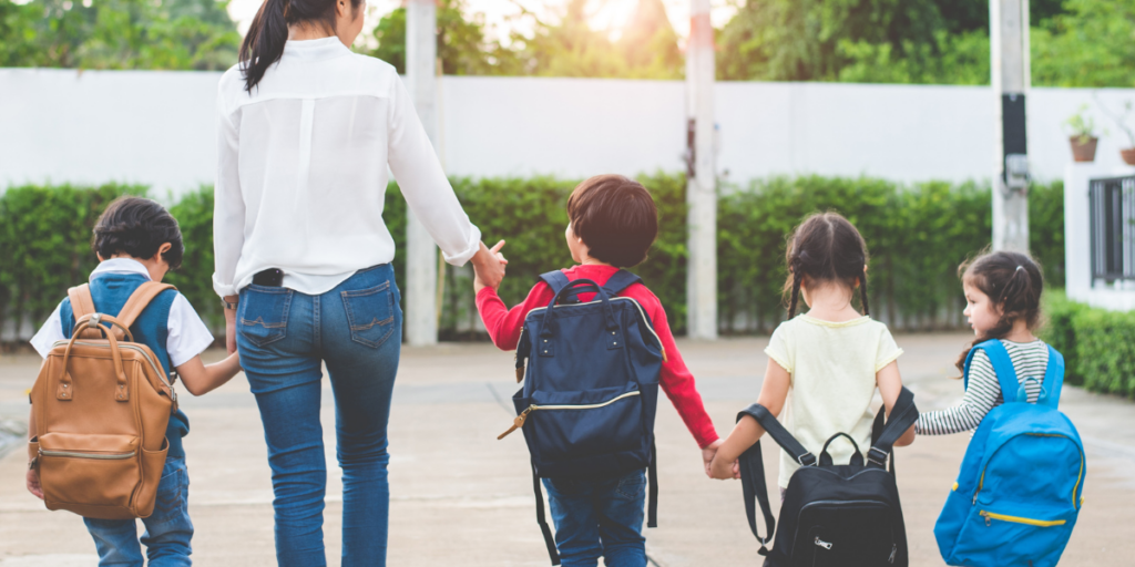 Four children with backpacks hold hands while walking with an adult woman.