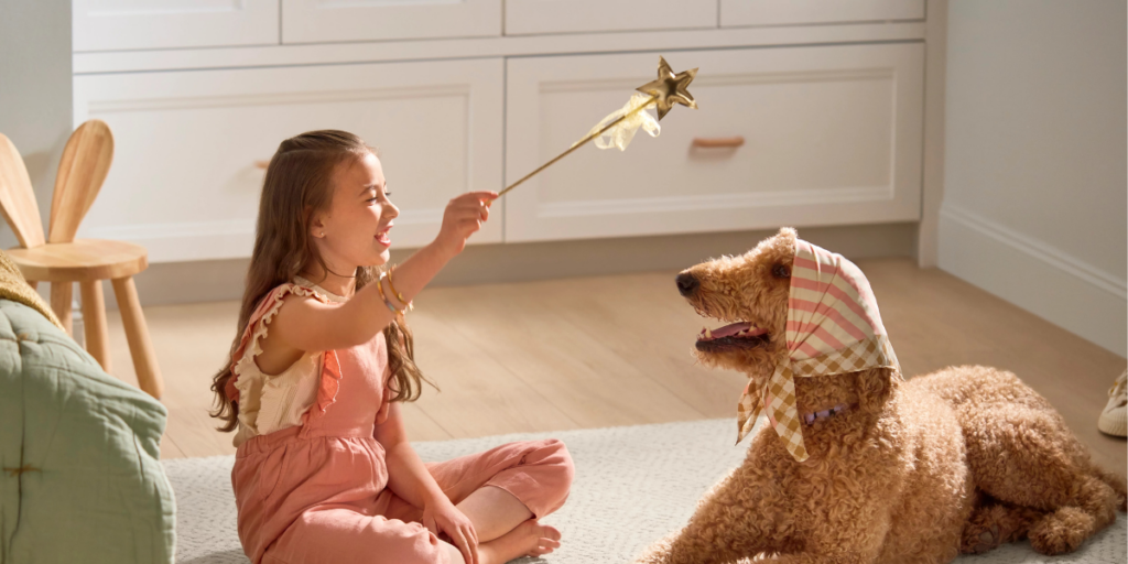 A young girl sits across from a large poodle. She is smiling and holding a toy magic wand that the dog is looking at.