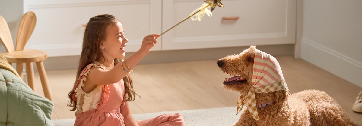 A young girl sits across from a large poodle. She is smiling and holding a toy magic wand that the dog is looking at.