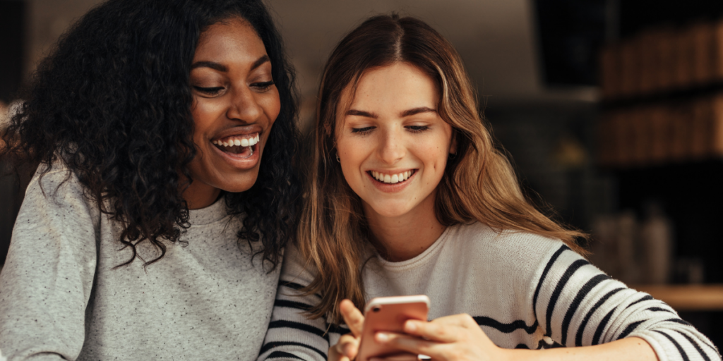 Two women sit next to each other, smiling and looking at a cellphone the one woman is holding.