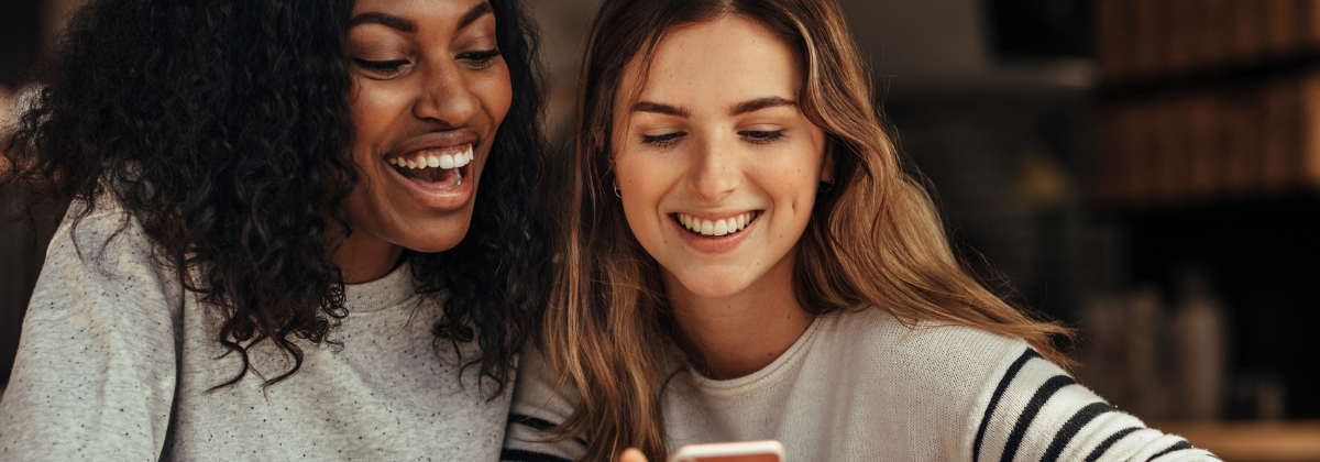 Two women sit next to each other, smiling and looking at a cellphone the one woman is holding.